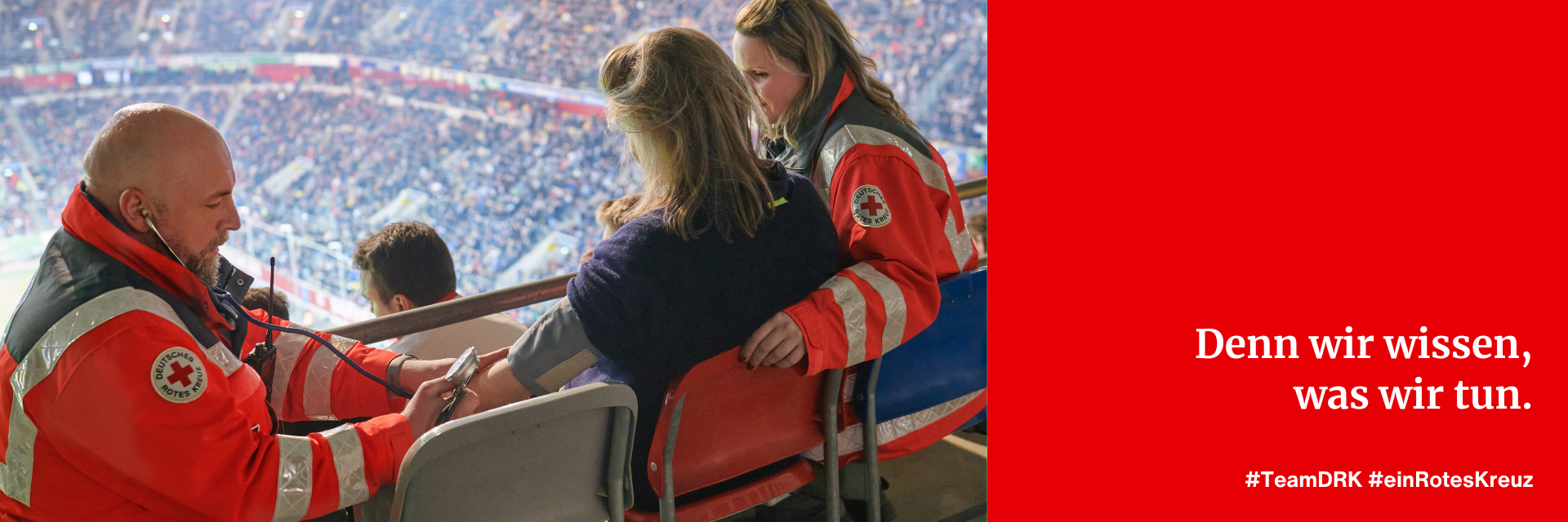 Eine männliche und eine weibliche Einsatzkraft des Deutschen Roten Kreuz versorgen eine Zuschauerin in einem Fußballstadion. Die männliche Einsatzkraft misst den Blutdruck der Zuschauerin.